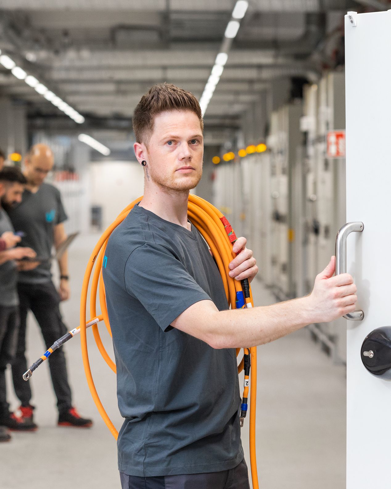 Ein Voltavision-Mitarbeiter im Voltavision-T-Shirt und mit einem orangefarbenen Kabel über der Schulter steht vor einer geöffneten Testkammer. Im Hintergrund sind drei weitere Mitarbeitende um einen Laptop geschart zu erkennnen.