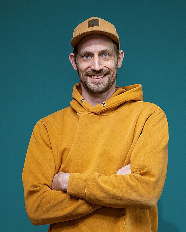 Eine Porträtaufnahme eines Voltavision-Mitarbeiters mit orangefarbener Cap und gleichfarbigem Kapuzenpullover vor petrolfarbenem Hintergrund.