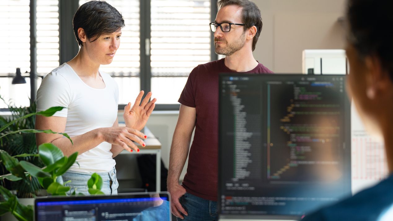 Drei Web-Entwickler:innen von Labor b im fachlichen Dialog. Im Bildvordergrund sind Screens mit Code zu sehen.