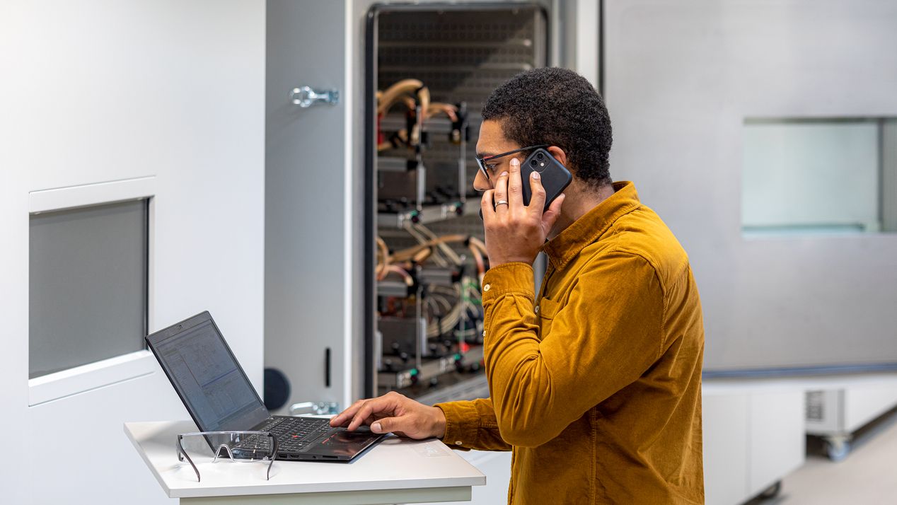Ein Voltavision-Mitarbeiter steht telefonierend an einem Stehpult und schaut auf einen Laptop. Im Hintergrund ist eine geöffnete Testkammer zu sehen.