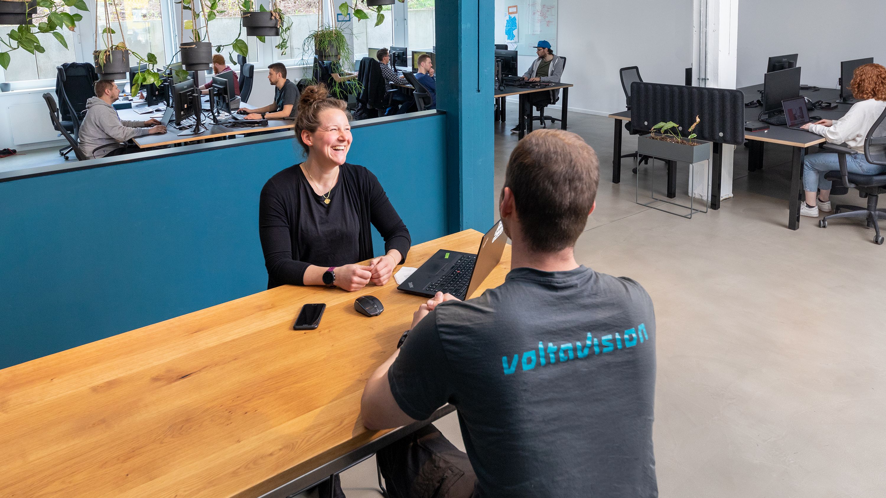 Ein Voltavision-Mitarbeiter im Voltavision-T-Shirt sitzt einer lachenden Mitarbeiterin gegenüber an einem Tisch in einem Großraumbüro. Im Hintergrund sind zahlreiche weitere Mitarbeitende an ihren Arbeitsplätzen zu sehen.