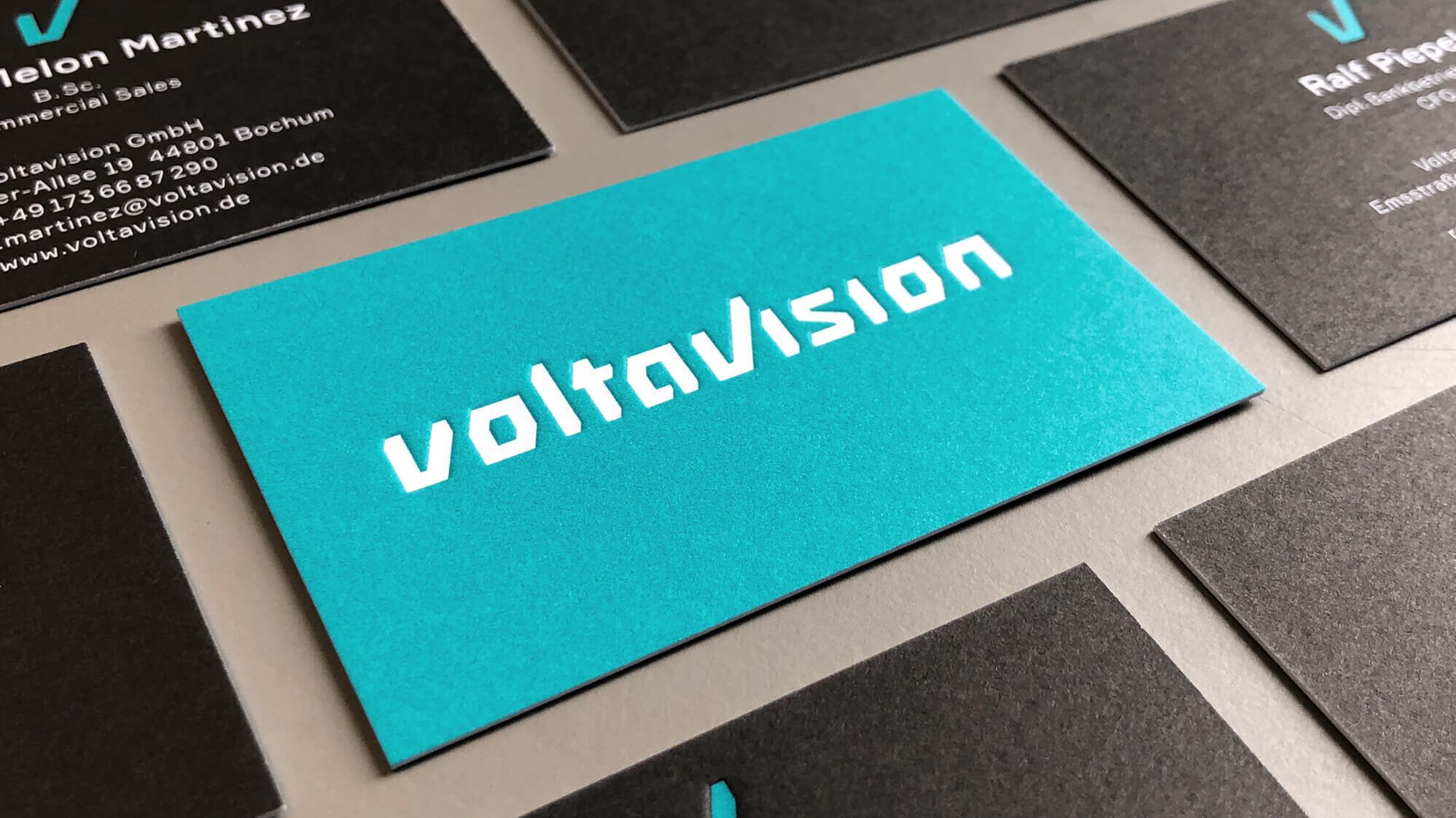 Die von Labor b gestalteten Voltavision-Visitenkarten mit geprägtem, weißen Logo-Schriftzug auf petrolfarbenem Grund auf der Vorderseite und anthrazitfarbener Rückseite mit Personennamen und Kontaktdaten.